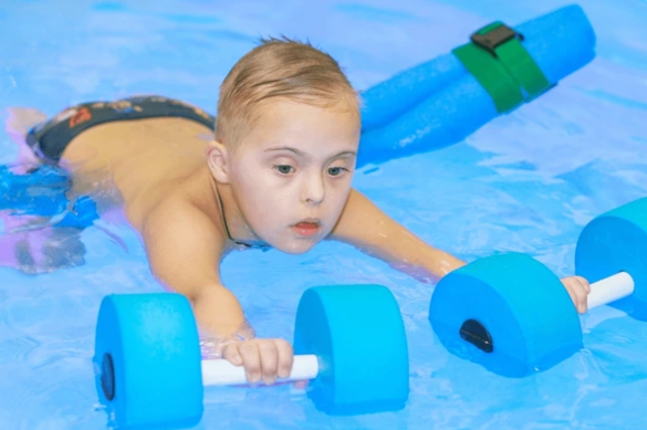 Picture1311 - Milestone CDC Young boy in a swimming pool, gripping a blue foam dumbbell with a white handle as he practices swimming in clear blue water.